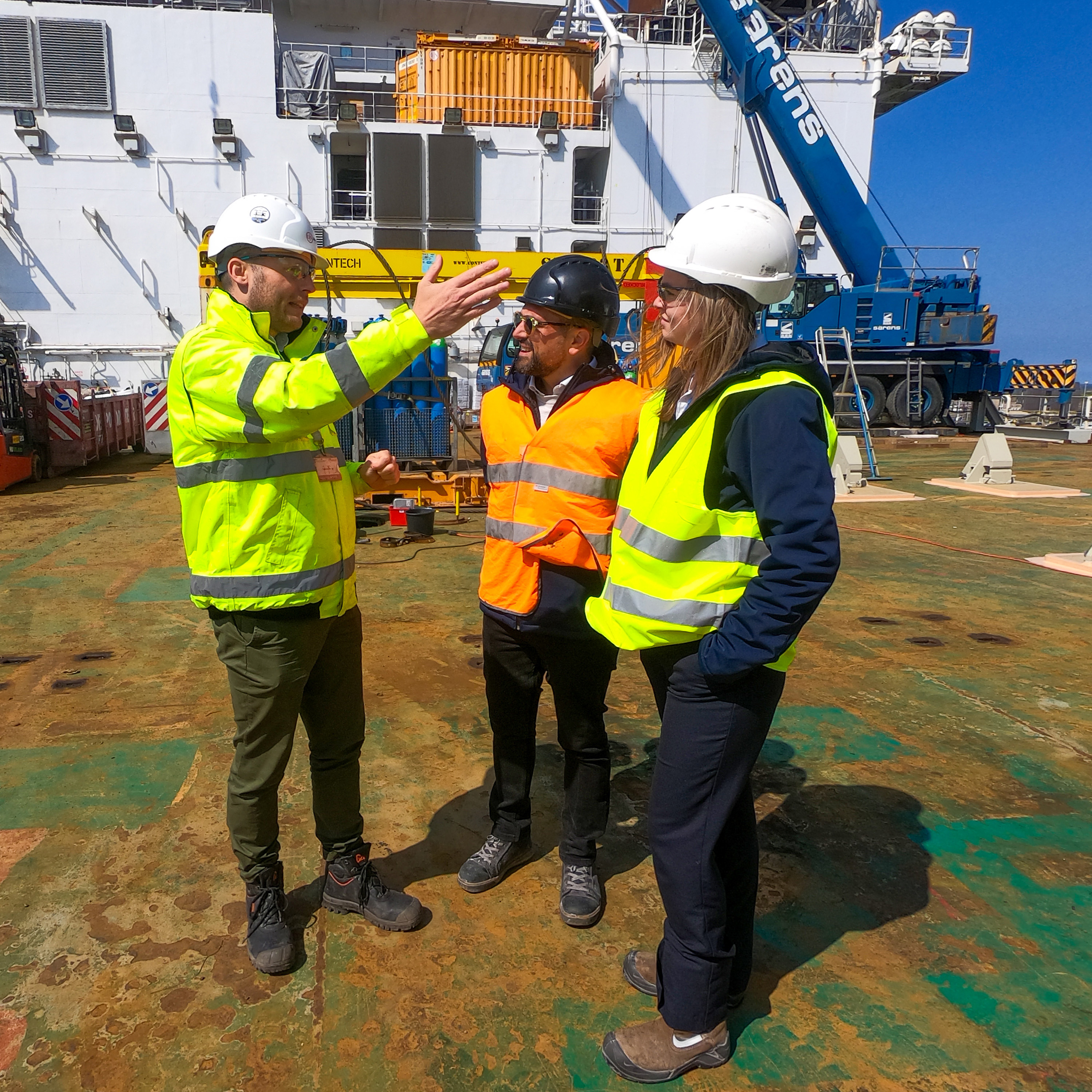 Lead Engineer in safety gear explains how technology works on the deck of an offshore vessel, with equipment, cranes, and clear blue sky in the background
